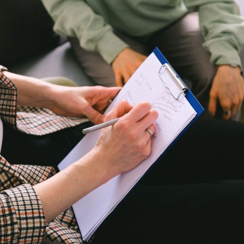 Crop anonymous female coach writing notes on paper in clipboard while talking with client in room